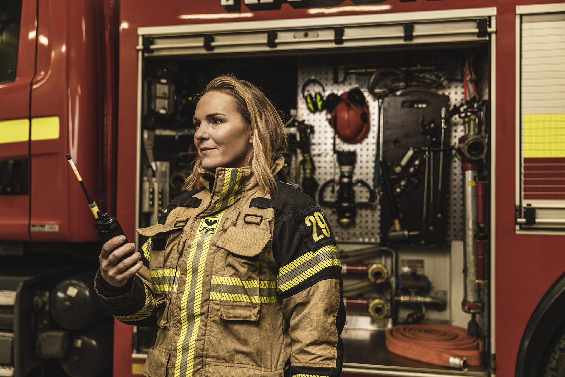 brandmanRIB En kvinnlig brandman i larmställ står framför en brandbil med en walkie-talkie i ena handen.