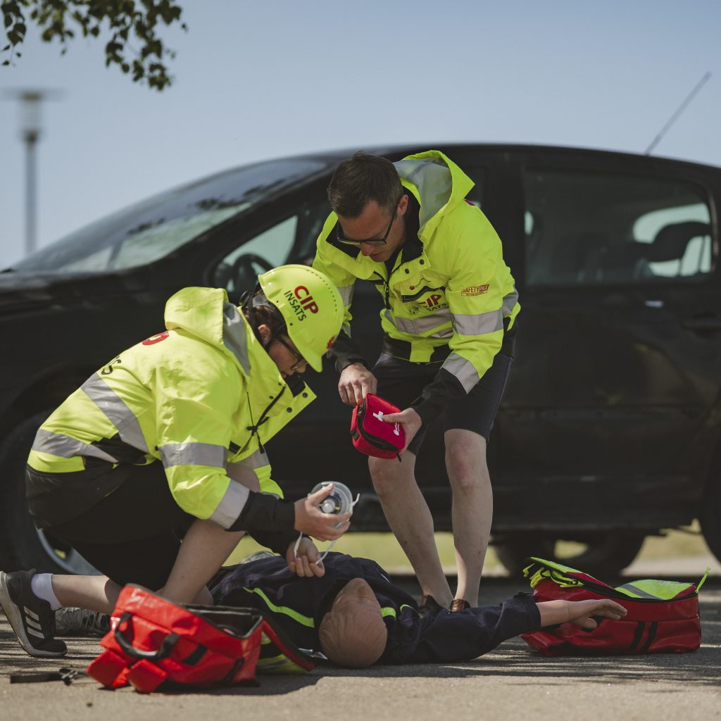 Två personer övar på att rädda en docka som ligger på marken framför en bil. Båda två är iklädda varselkläder där det står civil insatsperson på.
