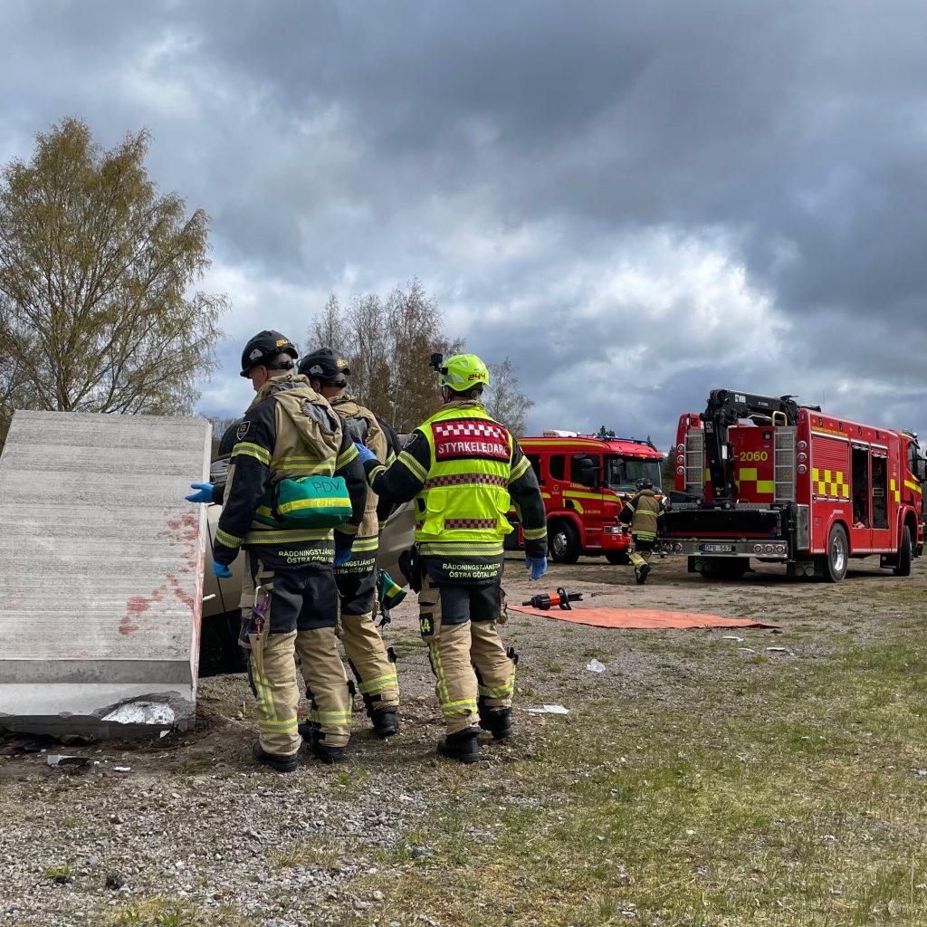 Räddningstjänstpersonal står intill ett stort fallet stenblock på en öppen plats. I bakgrunden står parkerade räddningsfordon.