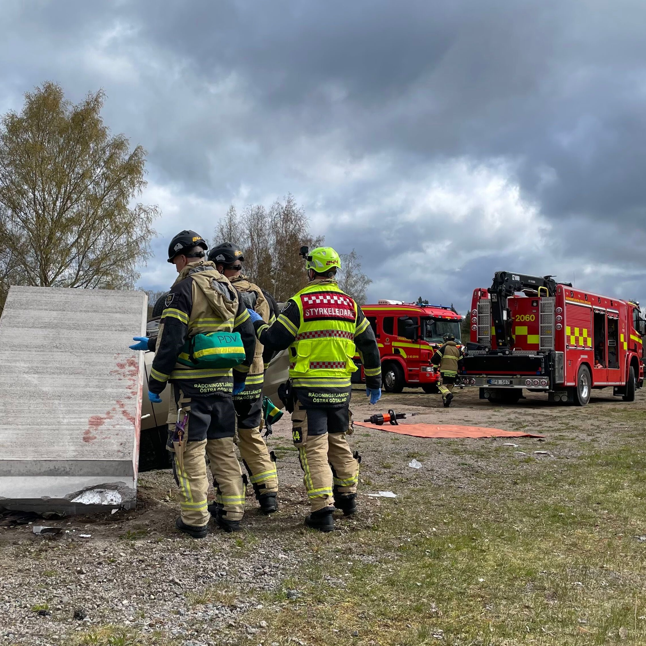 Räddningstjänstpersonal står intill ett stort fallet stenblock på en öppen plats. I bakgrunden står parkerade räddningsfordon.