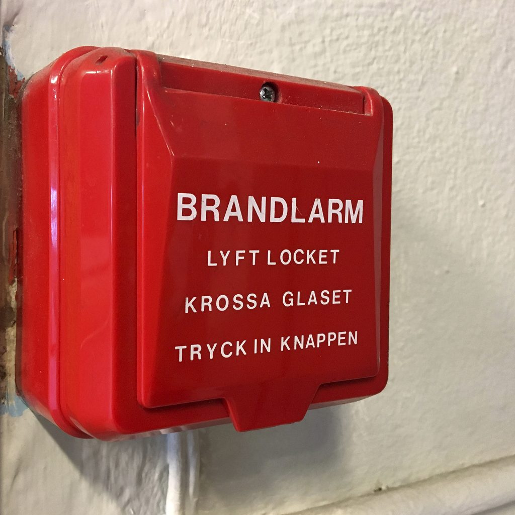 Ett automatlarm sitter monterat på en vägg. Det är en röd plastlåda och på står det "Brandlarm"