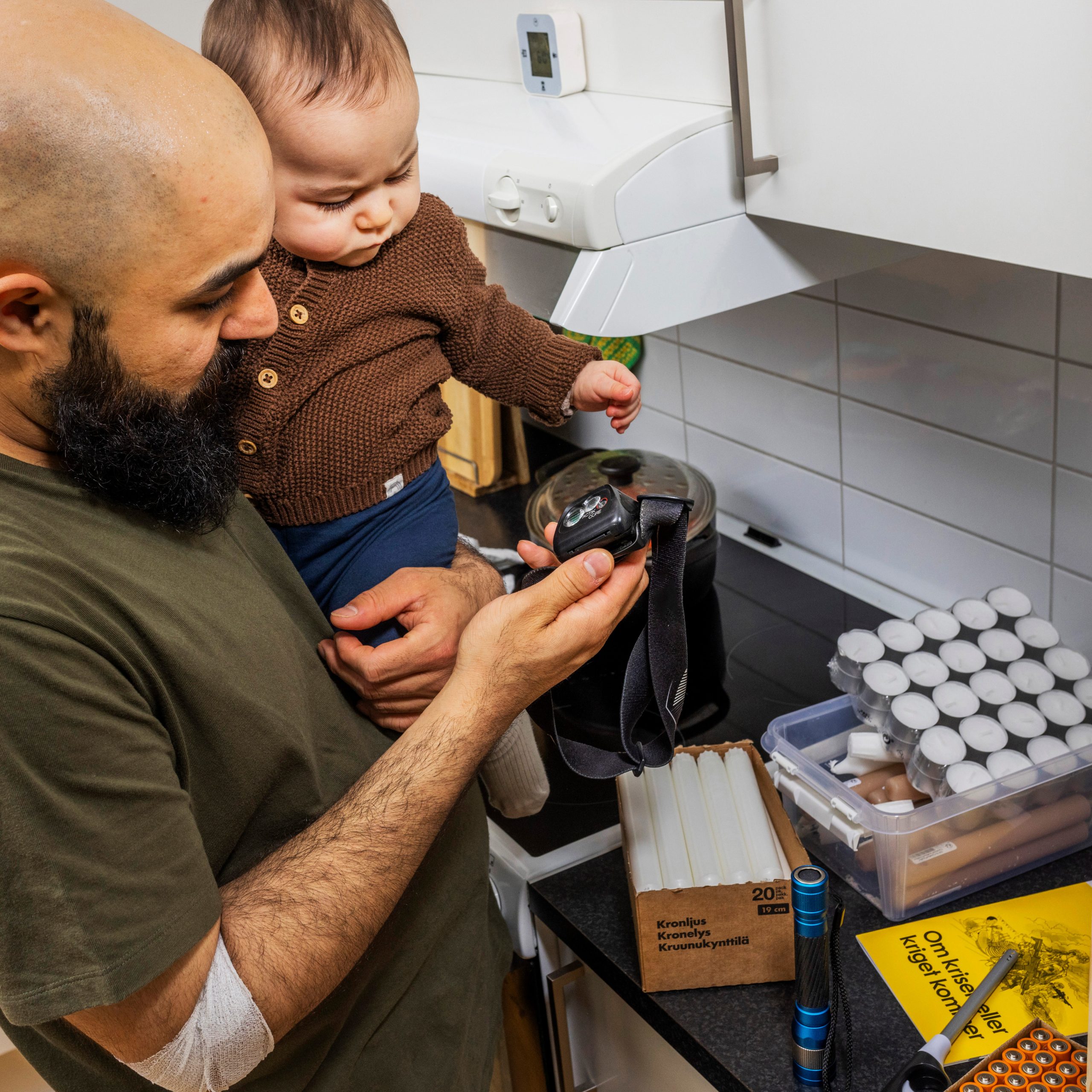 Bild på en man som håller ett barn på armen och tittar på en pannlampa. Framför honom står en låda med ljus och tändstickor på en köksbänk.