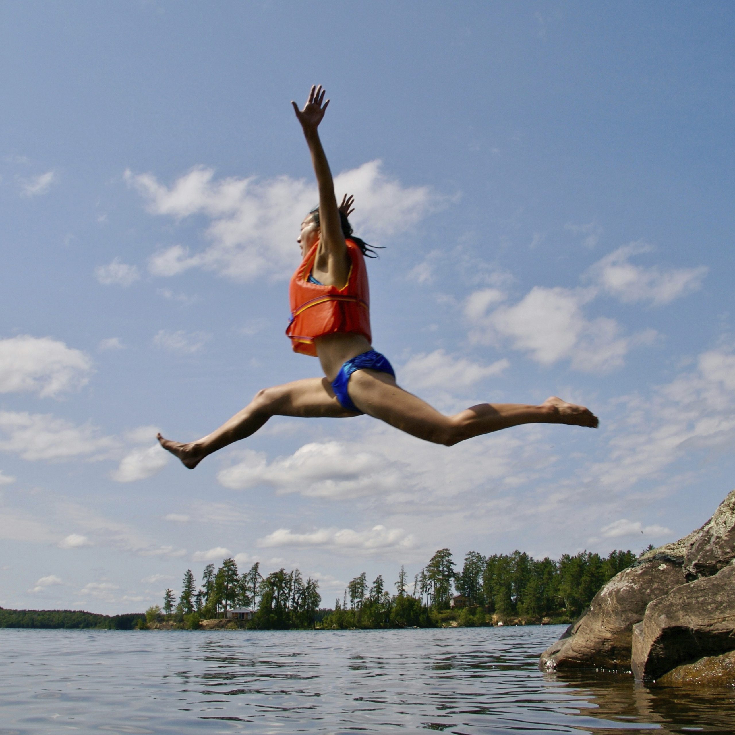 En kvinna gör ett stort hopp ut i vatten från en klippa. Hon har på sig en flytväst.