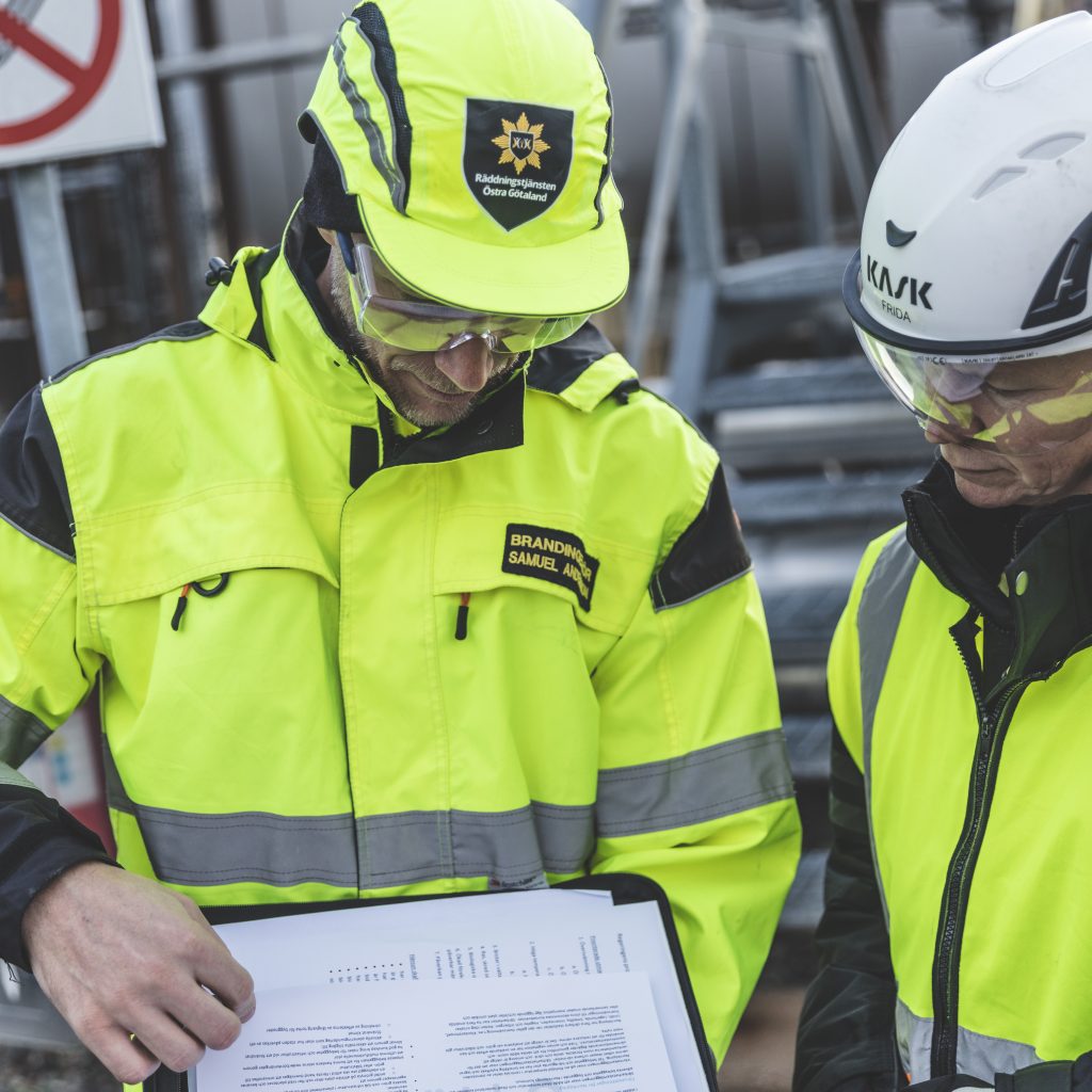 Två brandingenjörer iklädda varselkläder och hjälm står och tittat på dokumentationspapper.